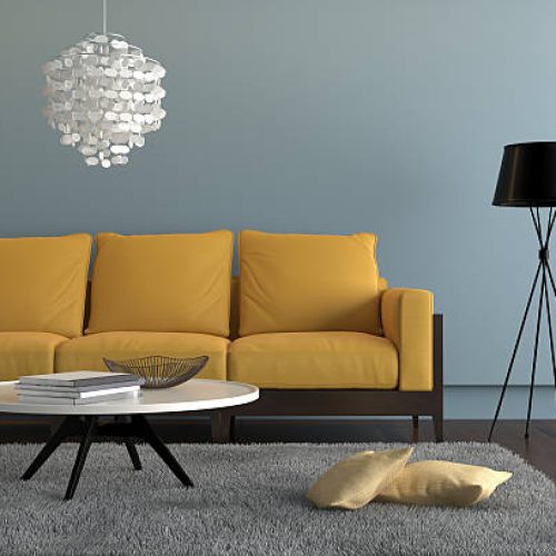 Close up view of a living room showing a yellow orange sofa, with a lamp, ornate pendant and a table with decoration. There is a gray carpet with two pillows on it. Light blue wall surrounding the scene with wooden floor. Horizontal composition.