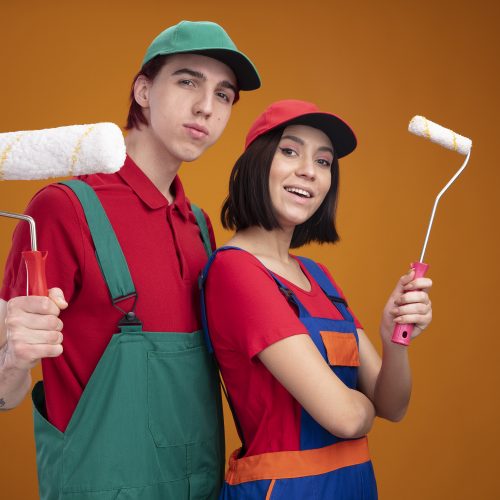 young couple confident guy and excited girl in construction worker uniform and cap standing in profile view holding paint roller looking at camera isolated on orange background